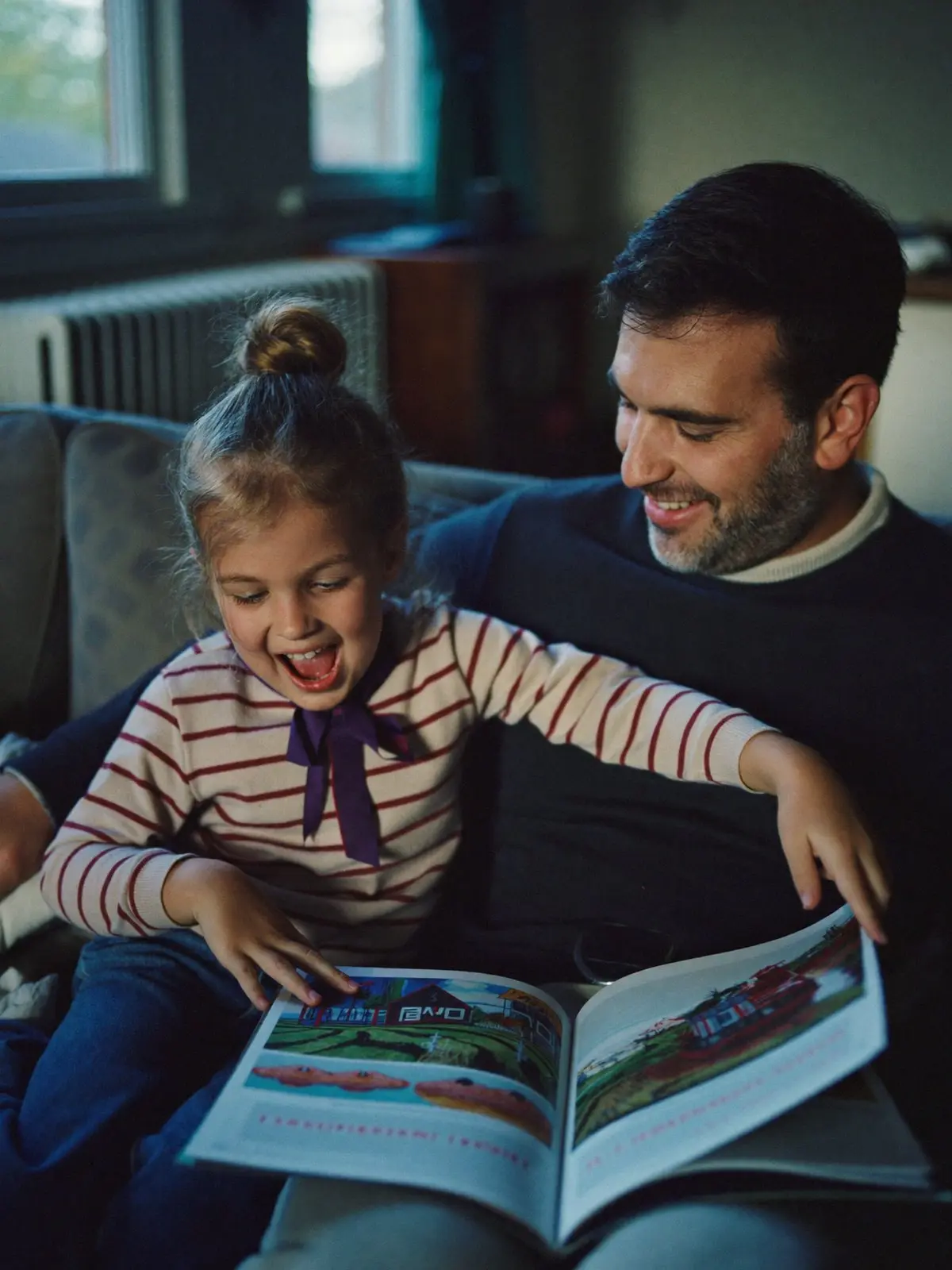 A father and his laughing daughter reading a picture book together on the sofa