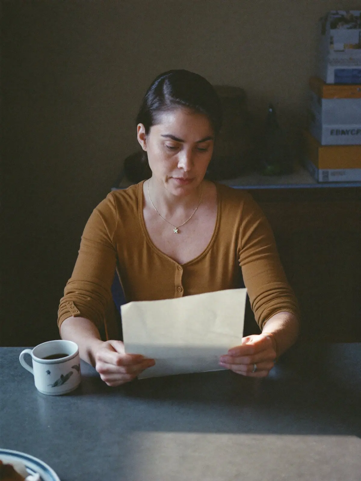 A woman at her kitchen table in the morning, reading a letter over a cup of coffee