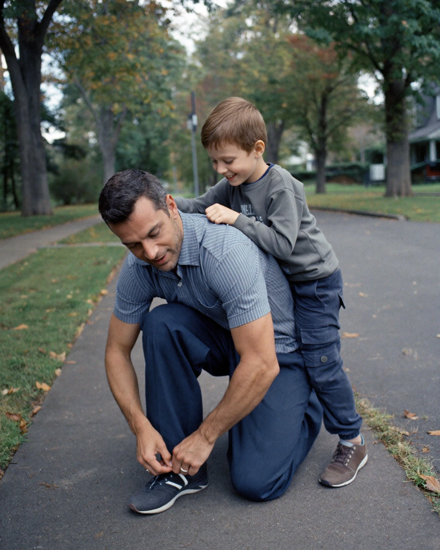 A father kneeling on a tree-lined sidewalk to tie his sneaker while his laughing son hangs on his back