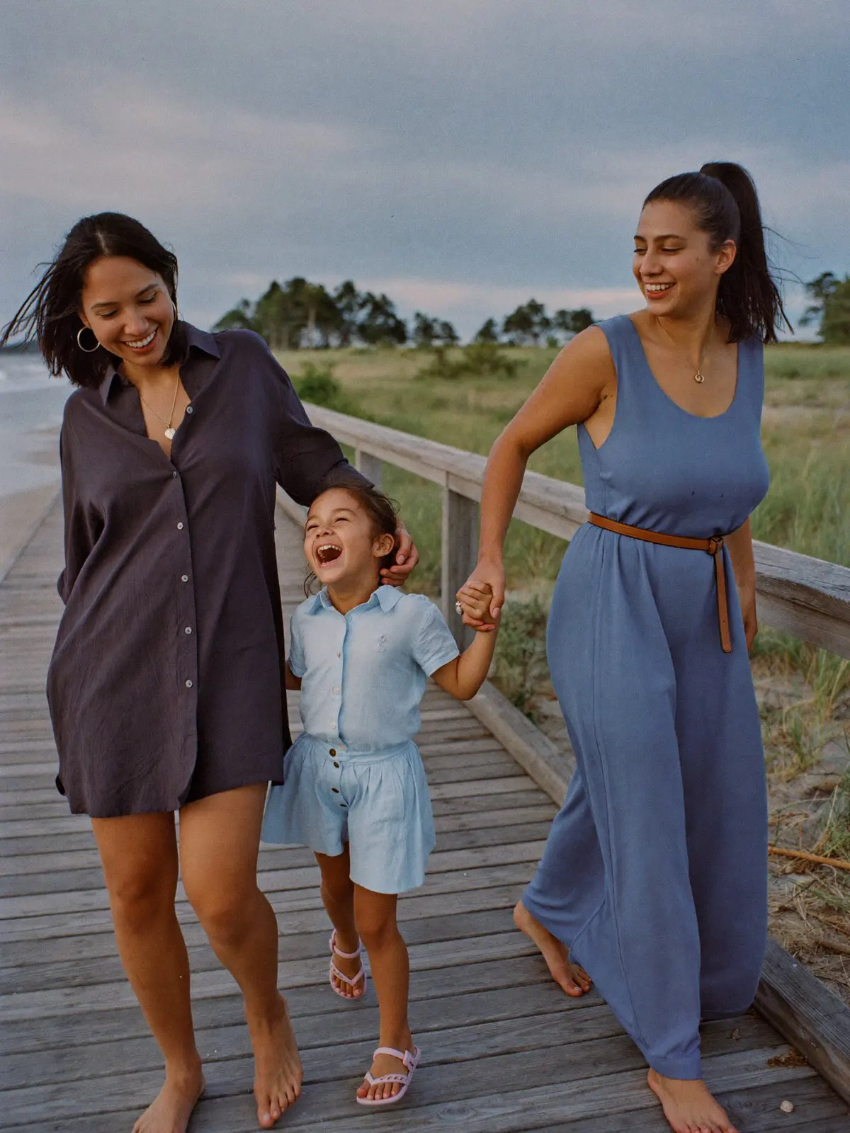 Two mothers walking their laughing daughter between them on a beach boardwalk